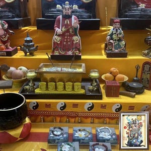 May include: A display of religious items, including statues, incense, and offerings, arranged on a yellow surface. The scene includes a framed artwork, small cups, and bracelets. The overall aesthetic suggests a cultural or spiritual context.