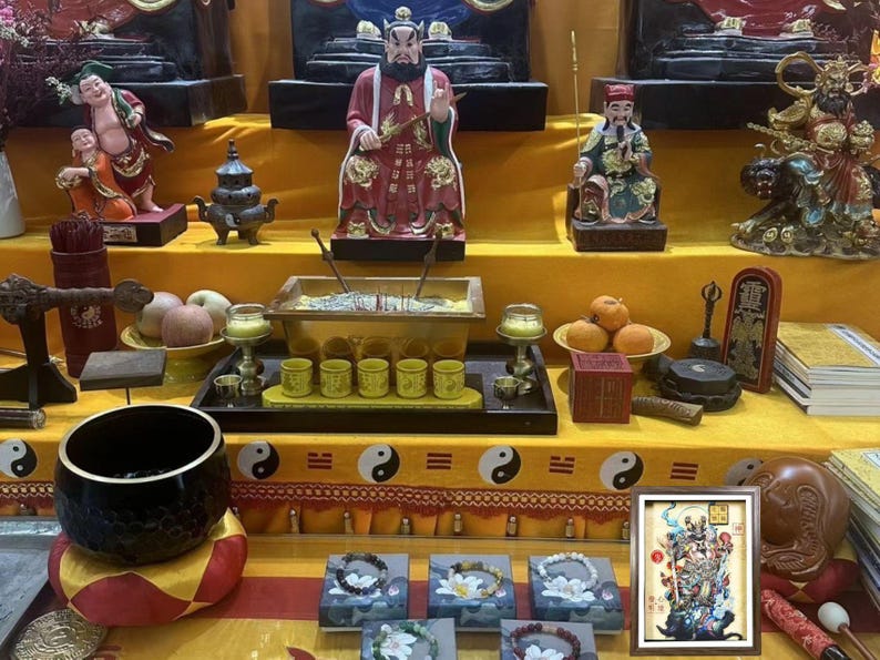May include: A display of religious items, including statues, incense, and offerings, arranged on a yellow surface. The scene includes a framed artwork, small cups, and bracelets. The overall aesthetic suggests a cultural or spiritual context.