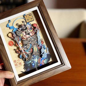 May include: A framed, multi-layered paper art piece featuring a detailed illustration of a deity or mythical figure. The artwork is predominantly colorful, with shades of blue, red, and gold. The frame is made of wood.