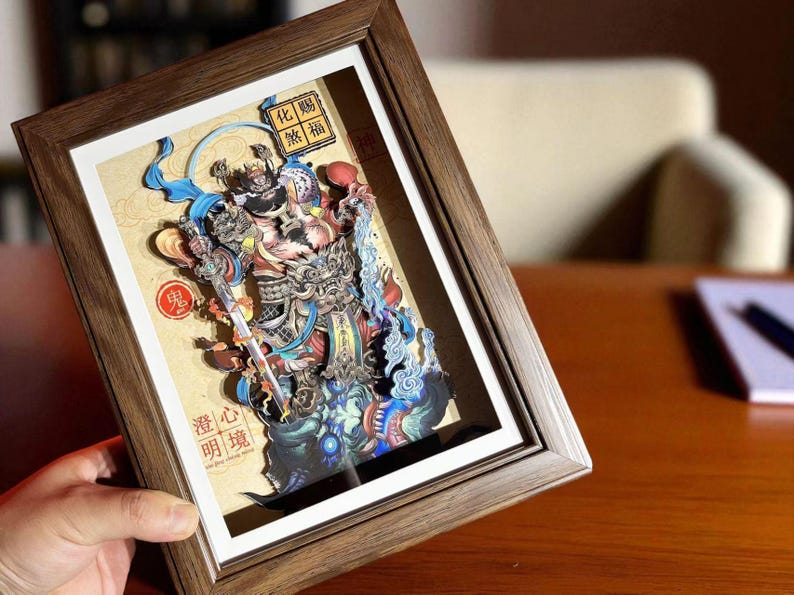 May include: A framed, multi-layered paper art piece featuring a detailed illustration of a deity or mythical figure. The artwork is predominantly colorful, with shades of blue, red, and gold. The frame is made of wood.