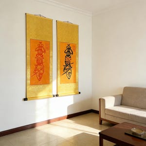 Puede incluir: Dos pinturas de desplazamiento asiáticas verticales cuelgan de una pared blanca. Cada rollo presenta un borde dorado, un centro naranja y caligrafía negra. Los rollos están montados con varillas negras y cuelgan cerca de un sofá beige.