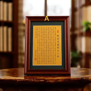 Könnte beinhalten: Ein gerahmtes Kunstwerk mit chinesischer Kalligraphie auf goldenem Hintergrund. Der Rahmen ist aus dunklem Holz, und das Kunstwerk wird auf einem Holztisch präsentiert. Der Text ist in Schwarz und Rot, mit dem Titel auf der rechten Seite.