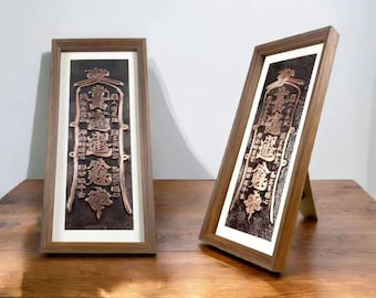 May include: Two framed copper-colored relief plaques with Asian calligraphy. The plaques are rectangular, with a dark background and a copper-colored design. The frames are made of wood. One plaque is leaning against a wooden stand.