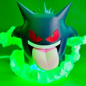 May include: A 3D-printed Gengar figure, a Pok&eacute;mon character, in dark blue with red eyes and a pink tongue. The figure is surrounded by green, glowing, cloud-like effects, set against a green background.