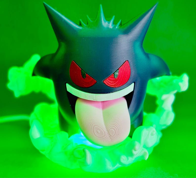 May include: A 3D-printed Gengar figure, a Pok&eacute;mon character, in dark blue with red eyes and a pink tongue. The figure is surrounded by green, glowing, cloud-like effects, set against a green background.