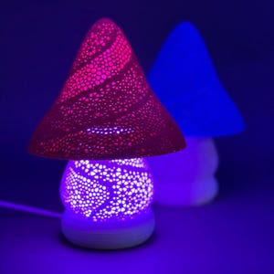 Pode incluir: Duas lâmpadas em forma de cogumelo. Uma tem uma sombra vermelha perfurada e uma base branca, emitindo um brilho roxo. A outra tem uma sombra azul. As lâmpadas estão em uma superfície azul escura. Decorativo.