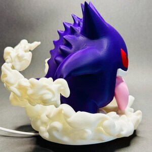 May include: A 3D-printed figurine of a purple creature with red eyes and a pink tongue, surrounded by white, cloud-like formations. The figurine has a spiky back and is set against a gray background.