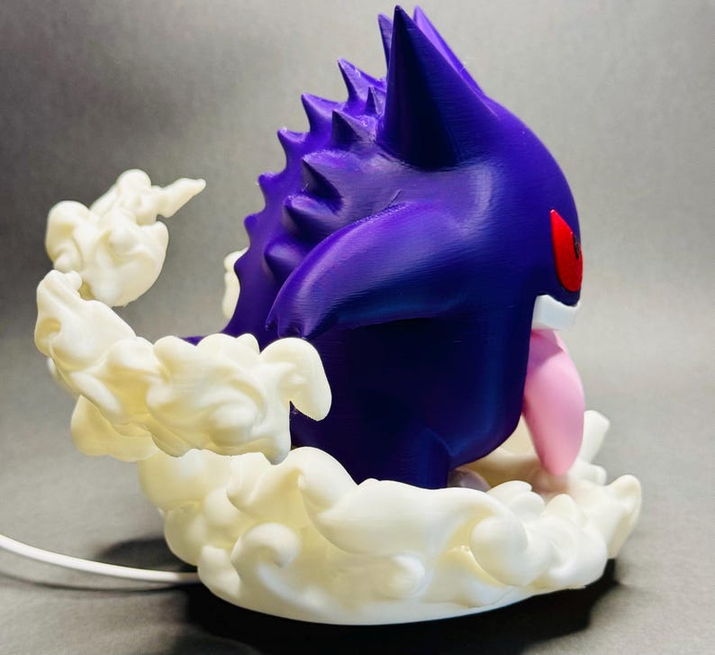 May include: A 3D-printed figurine of a purple creature with red eyes and a pink tongue, surrounded by white, cloud-like formations. The figurine has a spiky back and is set against a gray background.