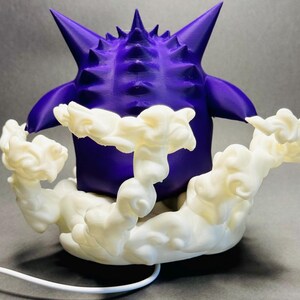 May include: A 3D-printed purple figure with a spiky head and arms, resting on a cloud-like base. The figure is a vibrant purple, and the cloud base is white. A white cord extends from the base.