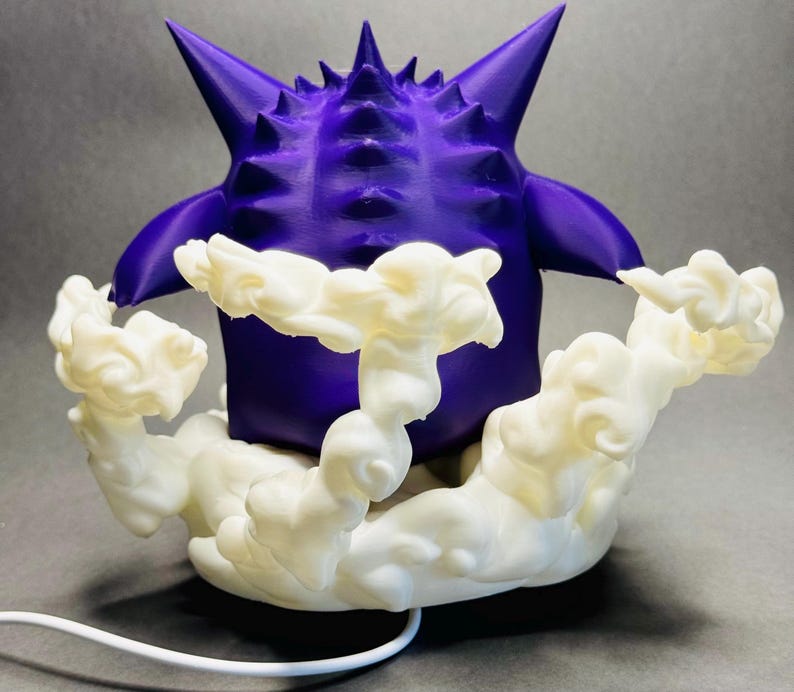May include: A 3D-printed purple figure with a spiky head and arms, resting on a cloud-like base. The figure is a vibrant purple, and the cloud base is white. A white cord extends from the base.