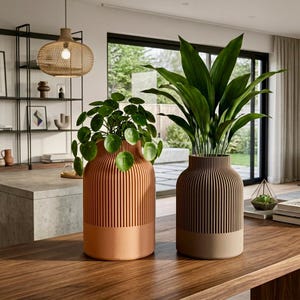 May include: Two cylindrical planters with vertical ribbed textures on the upper halves and smooth bases. The left planter is a light terracotta color containing a Pilea plant, while the right planter is a muted brown color holding a leafy green plant.