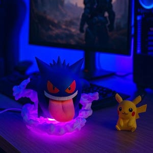 May include: A dark blue Gengar figurine with red eyes and a pink tongue, surrounded by a purple cloud-like base. A yellow Pikachu figurine sits nearby. The scene is illuminated with blue and purple lighting, suggesting a gaming or entertainment setup.