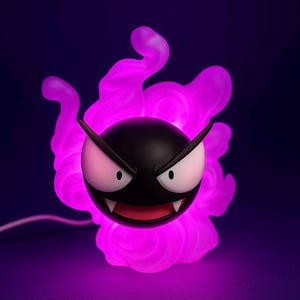 Puede incluir: Un personaje negro y redondo con ojos blancos, boca roja y dientes afilados, rodeado por una forma de nube púrpura brillante. El personaje tiene orejas puntiagudas y una expresión amenazante. Un cable blanco se extiende desde la base.
