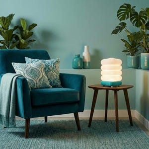 May include: A teal armchair with patterned cushions and a matching throw. A small wooden side table supports a white and teal lamp. Potted plants and vases are in the background. The room has a teal colour scheme.