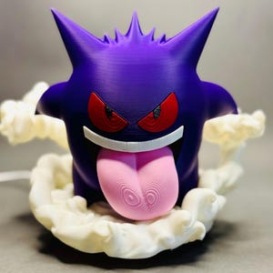 May include: A purple figurine of a cartoon character with red eyes, a wide grin, and a long pink tongue. The character is surrounded by a white, cloud-like base. The figurine has sharp, pointed ears and is likely a collectible item.