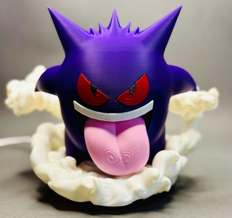May include: A purple figurine of a cartoon character with red eyes, a wide grin, and a long pink tongue. The character is surrounded by a white, cloud-like base. The figurine has sharp, pointed ears and is likely a collectible item.