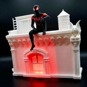 Puede incluir: Una fachada de edificio blanca impresa en 3D con una torre de agua en el techo, una escalera y una gárgola. Una figura de Spider-Man negra y roja está sentada en la cornisa. El área de la ventana está iluminada con una luz roja brillante.