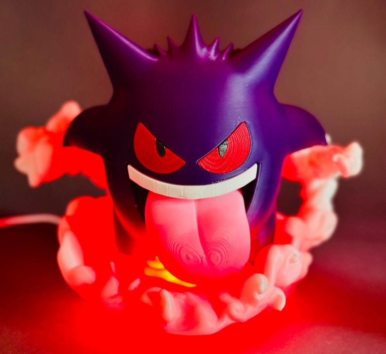 May include: A purple Gengar lamp with red eyes and a long pink tongue. The lamp is surrounded by a cloud of white smoke and emits a red glow. The design is based on the popular Pokemon character.