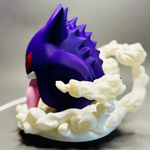 May include: A purple and white figurine of a Gengar character. The figurine has a purple body with red eyes and a pink tongue. It is surrounded by a white, cloud-like base.
