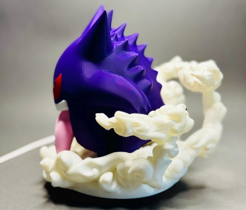 May include: A purple and white figurine of a Gengar character. The figurine has a purple body with red eyes and a pink tongue. It is surrounded by a white, cloud-like base.