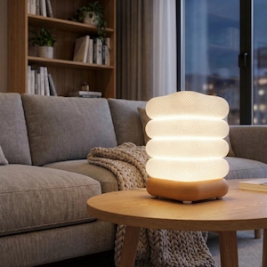May include: A modern, illuminated table lamp with a stacked, cylindrical design. The lamp has a warm, white glow and sits on a wooden base. The lamp is on a round wooden table in a living room setting.