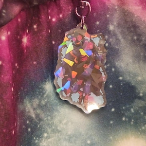 Peut inclure: Un porte-clés holographique avec un personnage, attaché à un fermoir argenté. Le charme est de forme irrégulière et reflète plusieurs couleurs. Le fond est un imprimé galaxie.