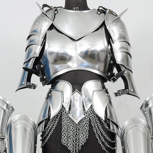 K&ouml;nnte beinhalten: Eine R&uuml;stung aus poliertem silberfarbenem Metall mit Brustpanzer, Schulterst&uuml;cken und Armschienen. Das Set enth&auml;lt Beinschienen und ein H&uuml;ftteil, das mit herabh&auml;ngenden silbernen Ketten verziert ist, pr&auml;sentiert auf einem schwarzen Torso.