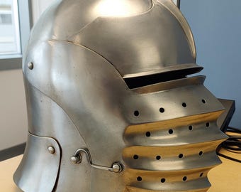 Könnte beinhalten: Ein polierter Schaller-Helm aus Stahl mit einer abgerundeten Helmglocke, einem horizontalen Sehschlitz und einem hervorstehenden Visier mit drei horizontalen Rippen, die kleine Belüftungslöcher enthalten. Der Helm hat einen seitlichen Haken und Nieten.