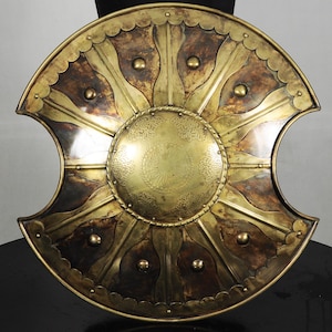 May include: A circular brass shield featuring a central embossed dome with intricate floral patterns. The shield has two deep crescent-shaped cutouts on the sides and is decorated with alternating dark brown panels and raised brass rivets.