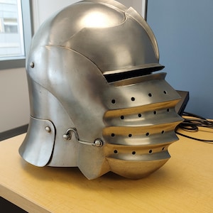 Puede incluir: Un casco medieval plateado con una parte superior redondeada y una visera con múltiples crestas horizontales y pequeños agujeros circulares. El casco tiene una placa para la mejilla con bisagras y se exhibe sobre una superficie de madera marrón claro.