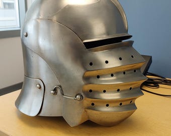 May include: A silver-colored medieval helmet with a rounded top and a visor featuring multiple horizontal ridges and small circular holes. The helmet has a hinged cheek plate and is displayed on a light brown wooden surface.
