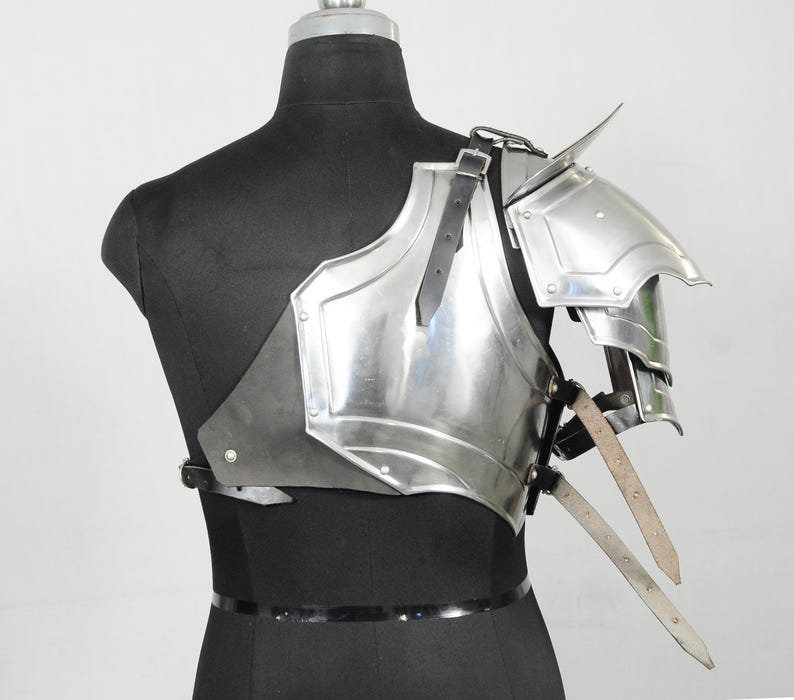 May include: A polished steel shoulder armour piece with a layered design, featuring a prominent shoulder spike. The armour is attached to a black leather backing and secured with leather straps. The piece is displayed on a black mannequin.