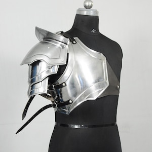 May include: A shiny, silver-coloured shoulder armour piece, possibly for cosplay or historical re-enactment. The armour has layered plates and is fastened with black leather straps and buckles. The item is displayed on a black mannequin.