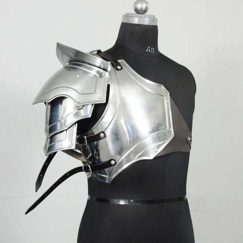 May include: A shiny, silver-coloured shoulder armour piece, possibly for cosplay or historical re-enactment. The armour has layered plates and is fastened with black leather straps and buckles. The item is displayed on a black mannequin.