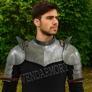 Könnte beinhalten: Ein silberfarbenes Schulterpanzer-Set aus Stahl mit braunen Lederriemen. Die Rüstung hat ein genietetes Design und wird über einem schwarzen Langarmshirt getragen. Der Text "VENDAROMY" ist auf dem Shirt aufgedruckt.