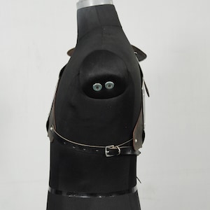 May include: A brown leather chest harness with silver metal accents and a buckle closure. The harness is displayed on a black mannequin, showcasing its design and construction. The leather appears to be distressed.