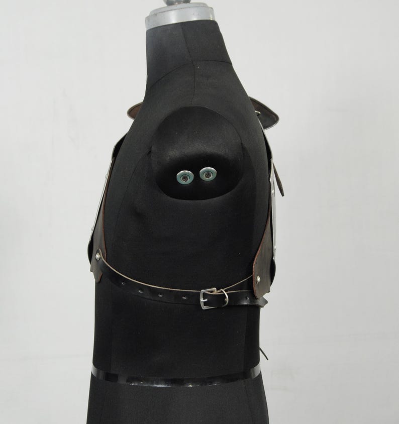 May include: A brown leather chest harness with silver metal accents and a buckle closure. The harness is displayed on a black mannequin, showcasing its design and construction. The leather appears to be distressed.