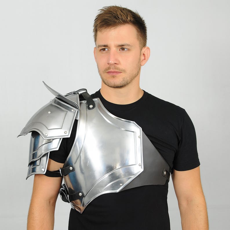 May include: A silver shoulder pauldron, a piece of armour, with layered plates and a curved design. The armour is attached to a black leather strap and is worn over a black t-shirt.