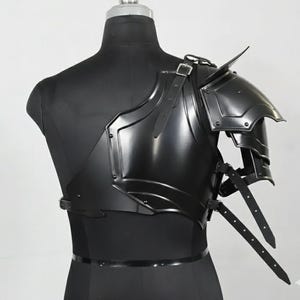 May include: Black metal shoulder armour with layered plates and leather straps. The armour is displayed on a black mannequin, showcasing its design and construction. The armour is designed to protect the shoulder and upper chest.