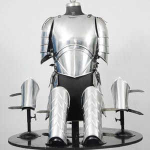 Könnte beinhalten: Ein komplettes Set aus polierter silberner mittelalterlicher Rüstung, einschließlich Brustpanzer, Schulterschutz, Arm- und Beinschutz. Die Rüstung wird auf einer schwarzen Schaufensterpuppe und einem runden schwarzen Tisch ausgestellt. Die Schaufensterpuppe hat die Nummer 10.