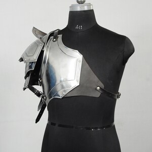 May include: A polished steel shoulder armour piece, displayed on a black mannequin. The armour features layered plates, rivets, and leather straps. The design is reminiscent of medieval or fantasy attire.