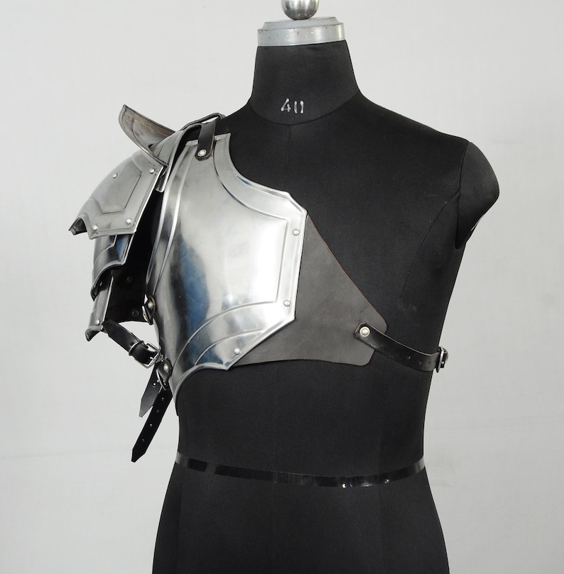May include: A polished steel shoulder armour piece, displayed on a black mannequin. The armour features layered plates, rivets, and leather straps. The design is reminiscent of medieval or fantasy attire.