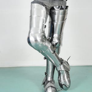 May include: Polished steel leg armor, featuring articulated plates for knee and shin protection. The armor includes attached foot coverings with pointed toes and is secured with leather straps and laces. This medieval-style armor is designed for historical reenactment or display.