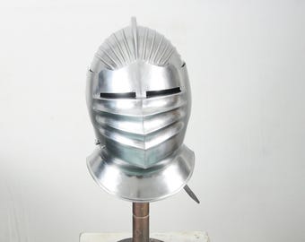 May include: A polished silver knight's helmet with a ridged top and a visor featuring horizontal eye slits. The helmet is mounted on a metal stand, showcasing its historical design and craftsmanship.