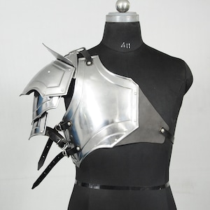 May include: A shiny silver shoulder armour piece with layered plates and a pointed edge. The armour is secured with black leather straps and buckles. The piece is displayed on a black mannequin.