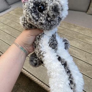 May include: A plush skunk toy made of soft, fuzzy white and mottled grey faux fur. The stuffed animal features a large, round black eye, a small pink crocheted flower detail on its head, and a long, bushy tail with a distinct white stripe.