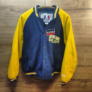 May include: A navy blue and yellow varsity jacket with the text "Levi's" and "Notre Dame" on the front. The jacket has a button-up closure, yellow sleeves, and a ribbed collar and cuffs. The jacket is hanging on a wooden hanger.