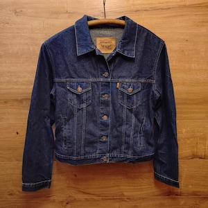 Op de afbeelding: Een donkerblauwe spijkerjas met een klassiek ontwerp. De jas heeft een knoopsluiting, twee borstzakken en een kraag. De jas hangt aan een houten kleerhanger tegen een houten achtergrond.