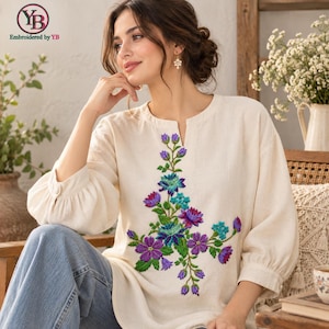 May include: A cream-colored long-sleeve blouse featuring a central vertical floral embroidery design in vibrant shades of purple, teal, and green. The garment has a simple round neckline with a small front slit and cuffed sleeves.