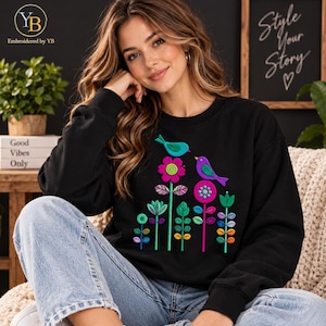 Pode incluir: Uma camisola preta de manga comprida com um design bordado de dois pássaros pousados em flores estilizadas coloridas. Os pássaros são azuis e roxos, enquanto as flores são apresentadas em tons vibrantes de rosa, verde e laranja.